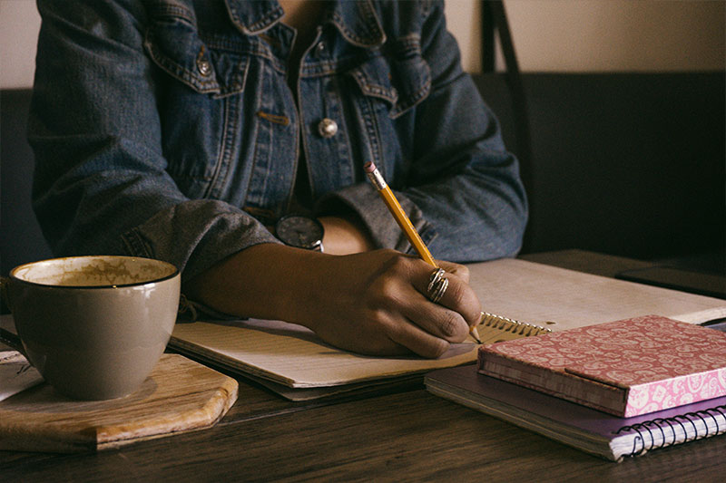 woman writing with a pencil in a notebook with a cup of coffee next to her